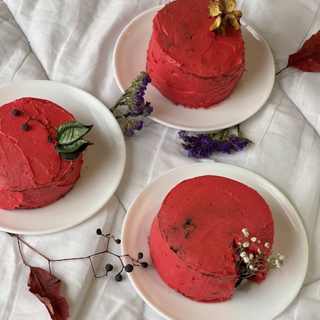 Three small, round cakes with vibrant red frosting are arranged on white plates. The cakes are garnished with various leaves and small decorative flowers. They are set on a white, slightly textured surface, creating a clean and minimalistic backdrop.