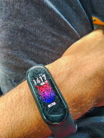 A close-up shot of a person's wrist wearing a smart fitness band. The band displays the time as 14:17, with a vibrant digital graphic of a tiger in red and blue hues. The screen shows signs of wear with visible scratches.