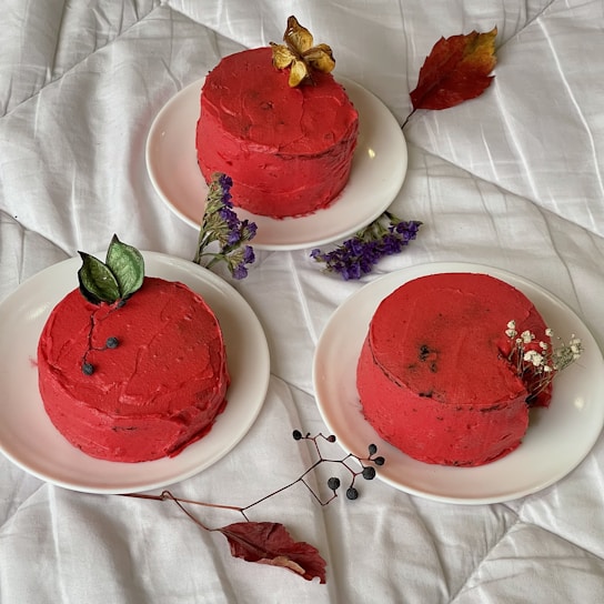Three small round cakes with red frosting are elegantly arranged on white plates. Each cake is decorated with different types of foliage and flowers, adding a touch of nature. The cakes are placed on a white textured surface, possibly a blanket, giving a cozy and artistic presentation. Nearby are scattered leaves and small branches for decoration.