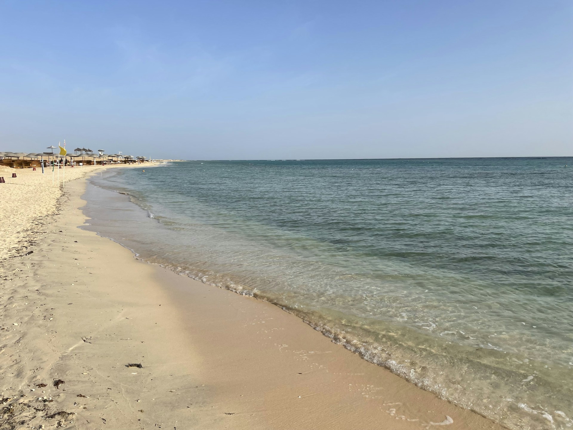A serene beach scene with soft white sand, gentle waves, and a glimpse of the Agio Portocervo apartment in the background.