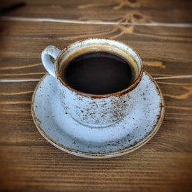 A rustic, speckled cup filled with dark coffee sits on a matching saucer against a textured wooden table. The cup and saucer have a worn look, featuring a white base with brown speckles.