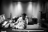 A person wearing a hat sits in a recording studio, adjusting equipment on a soundboard. Multiple screens display audio tracks, and musical instruments like a guitar are visible in the background. The room has an organized and functional appearance.