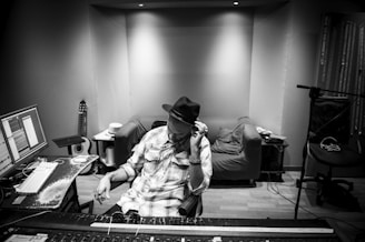 A person wearing a hat sits in a recording studio, adjusting equipment on a soundboard. Multiple screens display audio tracks, and musical instruments like a guitar are visible in the background. The room has an organized and functional appearance.