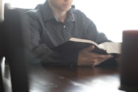 A person is sitting at a table, attentively reading an open book. The scene is softly lit, creating a serene and contemplative atmosphere. The focus is on the hands holding the book and the text they are engrossed in.