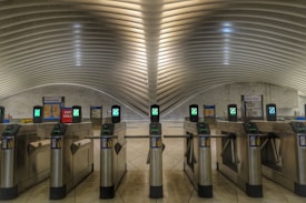 A modern transportation terminal with a series of turnstiles lined up under an architecturally designed ceiling with smooth, curved lines. The turnstiles have digital displays and card readers. Surrounding signs indicate exit only and closed platforms.