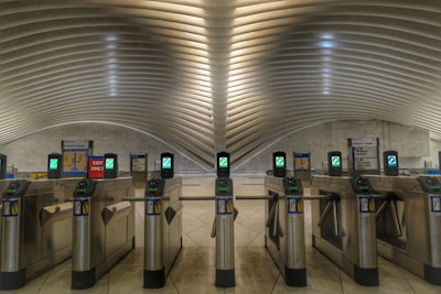 A modern transportation terminal with a series of turnstiles lined up under an architecturally designed ceiling with smooth, curved lines. The turnstiles have digital displays and card readers. Surrounding signs indicate exit only and closed platforms.