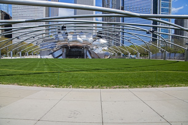 An outdoor venue with a large open grassy area surrounded by a modern, metallic structure that forms an arching canopy. Skyscrapers are visible in the background, adding an urban context to the scene. The sky is clear with a few clouds.