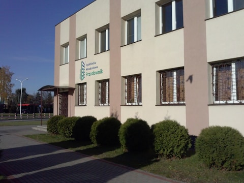 A modern, two-story building with multiple windows, a logo and text on the exterior wall. The surrounding area features trimmed shrubs and a paved walkway under a clear blue sky.