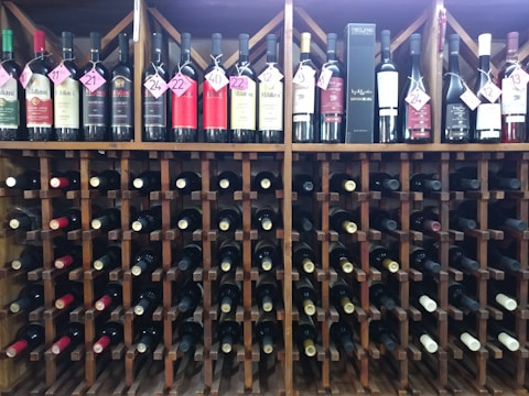 A wine rack filled with numerous bottles of wine. The bottles are organized neatly, some labeled with pink tags showing numbers. The structure of the rack is wooden, and the assortment includes various types of wine.