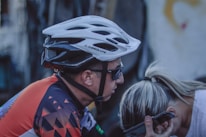 A cyclist wearing a white helmet and sunglasses appears to be talking on a smartphone while leaning towards another person with blonde hair. The cyclist is dressed in a colorful cycling jersey with geometric patterns, and the background is slightly blurred.