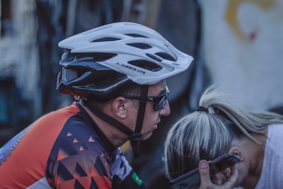 A cyclist wearing a white helmet and sunglasses appears to be talking on a smartphone while leaning towards another person with blonde hair. The cyclist is dressed in a colorful cycling jersey with geometric patterns, and the background is slightly blurred.