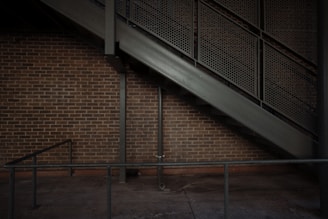 A brick wall is partially covered by a metal staircase with a perforated design. The stairs ascend diagonally from the left to the right, supported by vertical and diagonal metal beams. A simple metal railing runs parallel to the wall near the ground.