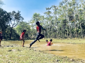 Children are playing near a muddy stream surrounded by trees. One child is mid-air, jumping with visible enthusiasm, while others are wading in the water. The scene is set in a natural environment with lush greenery.