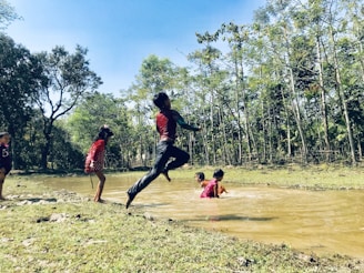 Children are playing near a muddy stream surrounded by trees. One child is mid-air, jumping with visible enthusiasm, while others are wading in the water. The scene is set in a natural environment with lush greenery.