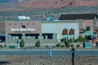 A building with signage for Black Bear Diner is situated in a desert landscape with rocky red hills in the background. The diner building has a beige and brown color scheme with stone accents and small bushes around its base. A road runs in front of the diner and a truck is visible in the background.