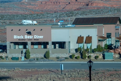 A building with signage for Black Bear Diner is situated in a desert landscape with rocky red hills in the background. The diner building has a beige and brown color scheme with stone accents and small bushes around its base. A road runs in front of the diner and a truck is visible in the background.