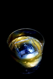 A glass with intricate diamond patterns filled with a light amber liquid, containing a large square ice cube. The lighting creates a dramatic contrast, highlighting the textures and clarity of the glass and ice against a dark background.