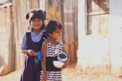 Two young children are standing outdoors with smiles on their faces. The child on the left is wearing a blue and white checked shirt with a dark purple pinafore, while the child on the right is dressed in a black and white striped dress and holding metallic containers. The background appears rustic, with blurred elements possibly indicating a rural setting.