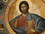 A religious icon depicting a figure in traditional attire holding a book with ornate designs, surrounded by Greek inscriptions. The background features circular golden and earthy tones.