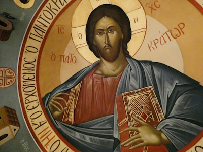 A religious icon depicting a figure in traditional attire holding a book with ornate designs, surrounded by Greek inscriptions. The background features circular golden and earthy tones.