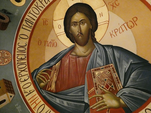 A religious icon depicting a figure in traditional attire holding a book with ornate designs, surrounded by Greek inscriptions. The background features circular golden and earthy tones.