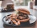 A juicy burger with a sesame seed bun, leafy lettuce, cheese, and various toppings sits on a dark plate. Crispy fries are scattered around the burger. Another burger is slightly blurred in the background, sitting on a similar plate.