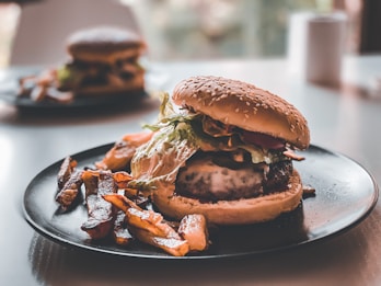A juicy burger with a sesame seed bun, leafy lettuce, cheese, and various toppings sits on a dark plate. Crispy fries are scattered around the burger. Another burger is slightly blurred in the background, sitting on a similar plate.