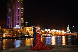 A couple is elegantly dressed and embracing beside a river at night, with illuminated modern buildings and city lights in the background. The woman is wearing a flowing red gown, and they appear affectionate and joyous in the serene, urban setting.