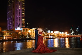 A couple is elegantly dressed and embracing beside a river at night, with illuminated modern buildings and city lights in the background. The woman is wearing a flowing red gown, and they appear affectionate and joyous in the serene, urban setting.
