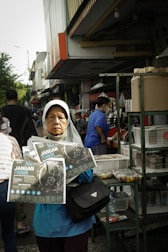 A woman wearing a headscarf stands in a busy outdoor market, holding pamphlets titled 'Jangan Panik.' She appears serious and focused. Behind her, people are shopping, and there are various stalls with goods displayed, including boxes and snacks.