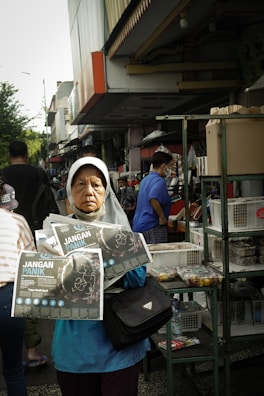 A woman wearing a headscarf stands in a busy outdoor market, holding pamphlets titled 'Jangan Panik.' She appears serious and focused. Behind her, people are shopping, and there are various stalls with goods displayed, including boxes and snacks.