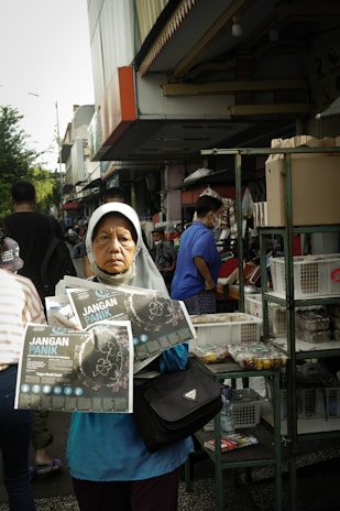 A woman wearing a headscarf stands in a busy outdoor market, holding pamphlets titled 'Jangan Panik.' She appears serious and focused. Behind her, people are shopping, and there are various stalls with goods displayed, including boxes and snacks.