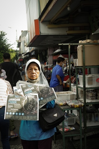 A woman wearing a headscarf stands in a busy outdoor market, holding pamphlets titled 'Jangan Panik.' She appears serious and focused. Behind her, people are shopping, and there are various stalls with goods displayed, including boxes and snacks.