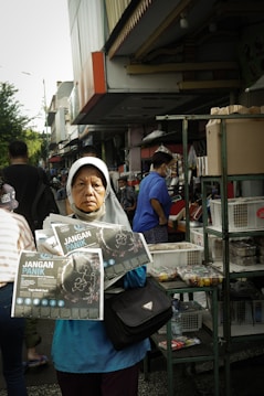 A woman wearing a headscarf stands in a busy outdoor market, holding pamphlets titled 'Jangan Panik.' She appears serious and focused. Behind her, people are shopping, and there are various stalls with goods displayed, including boxes and snacks.