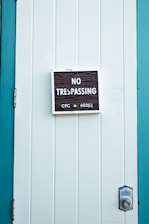 A close-up of a white paneled door with a 'No Trespassing' sign mounted in the center. The sign includes the text 'CPC sec 602(L)' in smaller print. The door is bordered by teal-colored framing, and a silver door latch is visible on the right side.