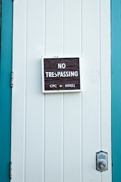 A close-up of a white paneled door with a 'No Trespassing' sign mounted in the center. The sign includes the text 'CPC sec 602(L)' in smaller print. The door is bordered by teal-colored framing, and a silver door latch is visible on the right side.