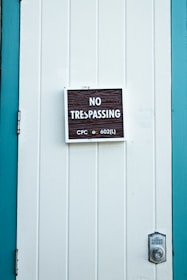 A close-up of a white paneled door with a 'No Trespassing' sign mounted in the center. The sign includes the text 'CPC sec 602(L)' in smaller print. The door is bordered by teal-colored framing, and a silver door latch is visible on the right side.