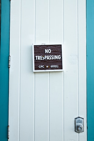 A close-up of a white paneled door with a 'No Trespassing' sign mounted in the center. The sign includes the text 'CPC sec 602(L)' in smaller print. The door is bordered by teal-colored framing, and a silver door latch is visible on the right side.
