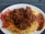 A plate of flavorful rice is topped with a rich, brown stew, likely containing pieces of meat. To the side, there is a serving of vibrant fresh salad, including sliced tomatoes and green vegetables, and a portion of red sauce.