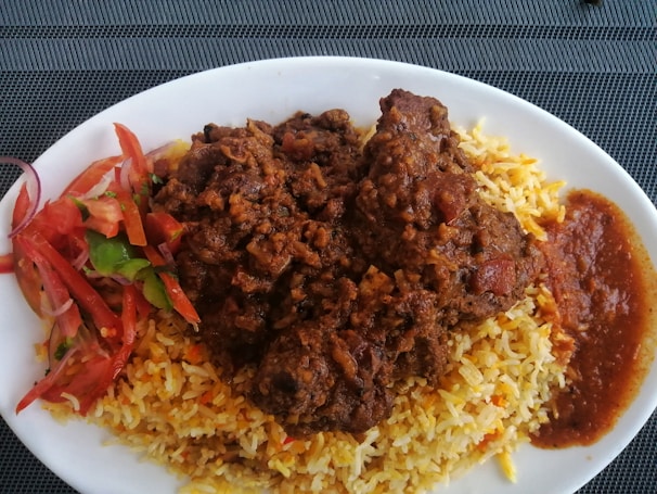 A plate of flavorful rice is topped with a rich, brown stew, likely containing pieces of meat. To the side, there is a serving of vibrant fresh salad, including sliced tomatoes and green vegetables, and a portion of red sauce.