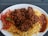 A plate of flavorful rice is topped with a rich, brown stew, likely containing pieces of meat. To the side, there is a serving of vibrant fresh salad, including sliced tomatoes and green vegetables, and a portion of red sauce.