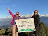 Two people are standing on a mountain with snow-capped peaks in the background, holding a banner that reads 'Heaven Himalaya Poonhill Treks 3210m'. Both are wearing colorful winter clothing, and one person is making a peace sign with their hand.