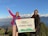 Two people are standing on a mountain with snow-capped peaks in the background, holding a banner that reads 'Heaven Himalaya Poonhill Treks 3210m'. Both are wearing colorful winter clothing, and one person is making a peace sign with their hand.