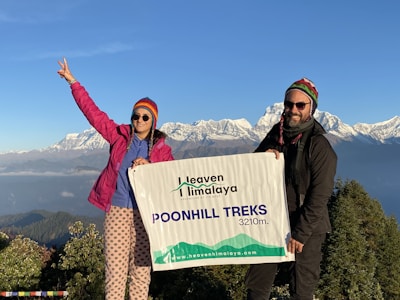 Two people are standing on a mountain with snow-capped peaks in the background, holding a banner that reads 'Heaven Himalaya Poonhill Treks 3210m'. Both are wearing colorful winter clothing, and one person is making a peace sign with their hand.