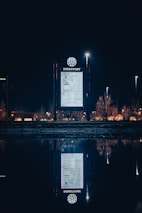 An illuminated directory sign is placed outdoors against a night sky. The sign is brightly lit and shows a map or directory layout. Surrounding it in the distance are lights from street lamps and buildings, with their reflections visible on a body of water in the foreground.
