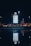 An illuminated directory sign is placed outdoors against a night sky. The sign is brightly lit and shows a map or directory layout. Surrounding it in the distance are lights from street lamps and buildings, with their reflections visible on a body of water in the foreground.