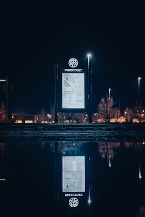 An illuminated directory sign is placed outdoors against a night sky. The sign is brightly lit and shows a map or directory layout. Surrounding it in the distance are lights from street lamps and buildings, with their reflections visible on a body of water in the foreground.