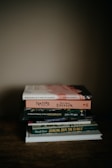 A stack of six books is piled neatly on a wooden surface. The books are varied in size and cover design, with titles including 'Birdsong in a Time of Silence' by Steven Lovatt and 'Nature' by Ralph Waldo Emerson. The scene is softly lit, creating a warm and cozy atmosphere.