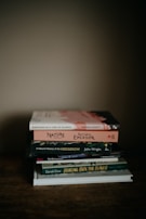 A stack of six books is piled neatly on a wooden surface. The books are varied in size and cover design, with titles including 'Birdsong in a Time of Silence' by Steven Lovatt and 'Nature' by Ralph Waldo Emerson. The scene is softly lit, creating a warm and cozy atmosphere.