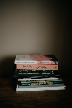 A stack of six books is piled neatly on a wooden surface. The books are varied in size and cover design, with titles including 'Birdsong in a Time of Silence' by Steven Lovatt and 'Nature' by Ralph Waldo Emerson. The scene is softly lit, creating a warm and cozy atmosphere.